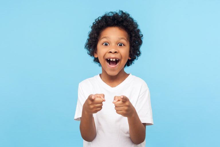 Portrait of happy little boy with curly hair pointing fingers to camera and laughing with surprised face