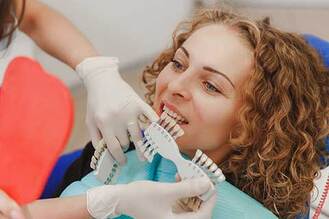 Patient seated in dental chair while dental professional wearing gloves holds shade guide with multiple tooth color samples near mouth to compare for cosmetic procedure.